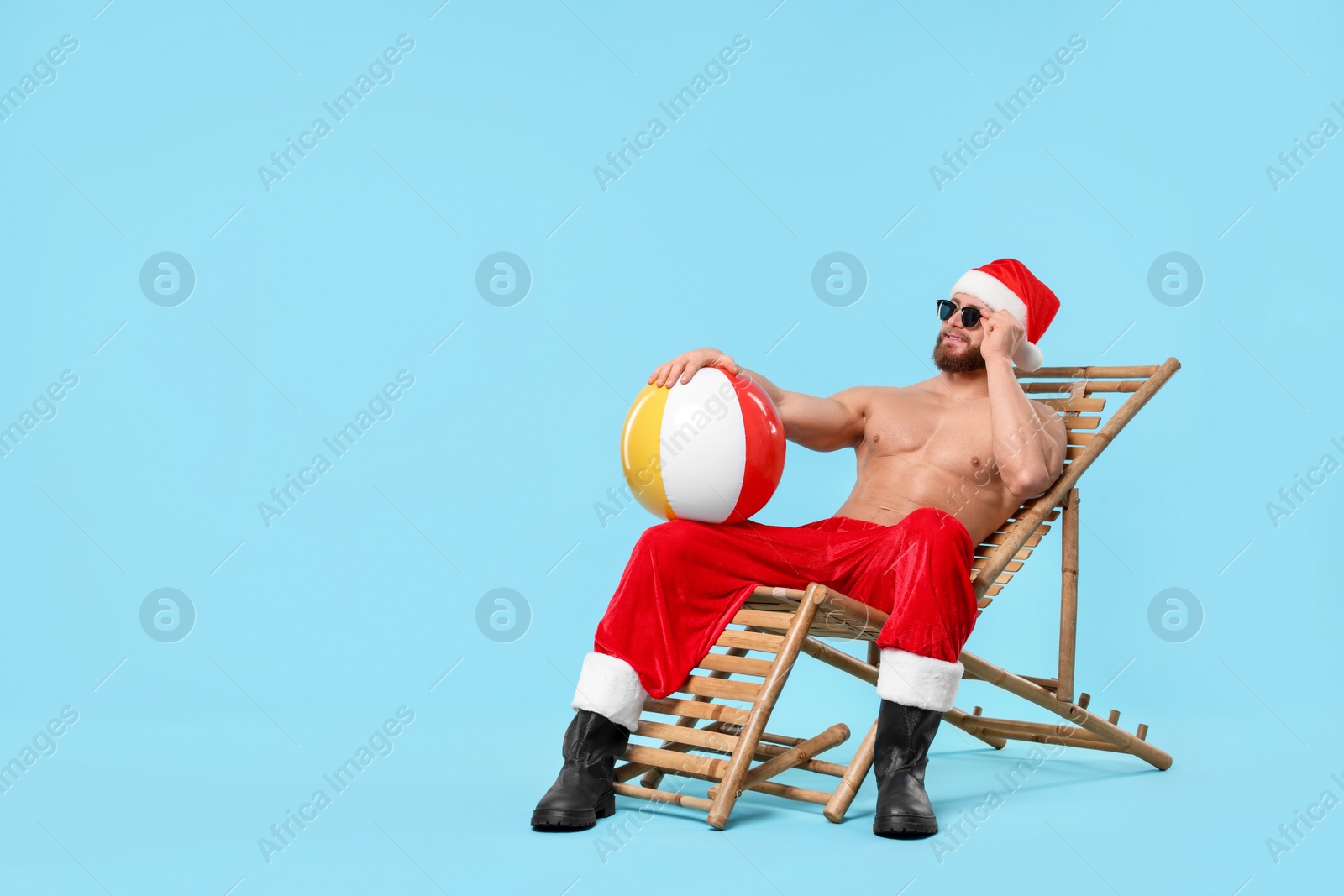 Photo of Muscular young man in Santa hat with ball, sunglasses and deck chair on light blue background, space for text
