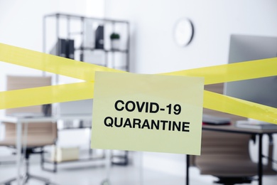 Photo of Sheet of paper with words COVID-19 Quarantine on crossed yellow tape in office