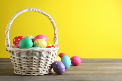 Photo of Colorful Easter eggs in wicker basket on wooden table against yellow background. Space for text