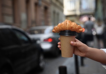 Photo of Woman holding tasty croissant and cup of coffee on city street, closeup. Space for text