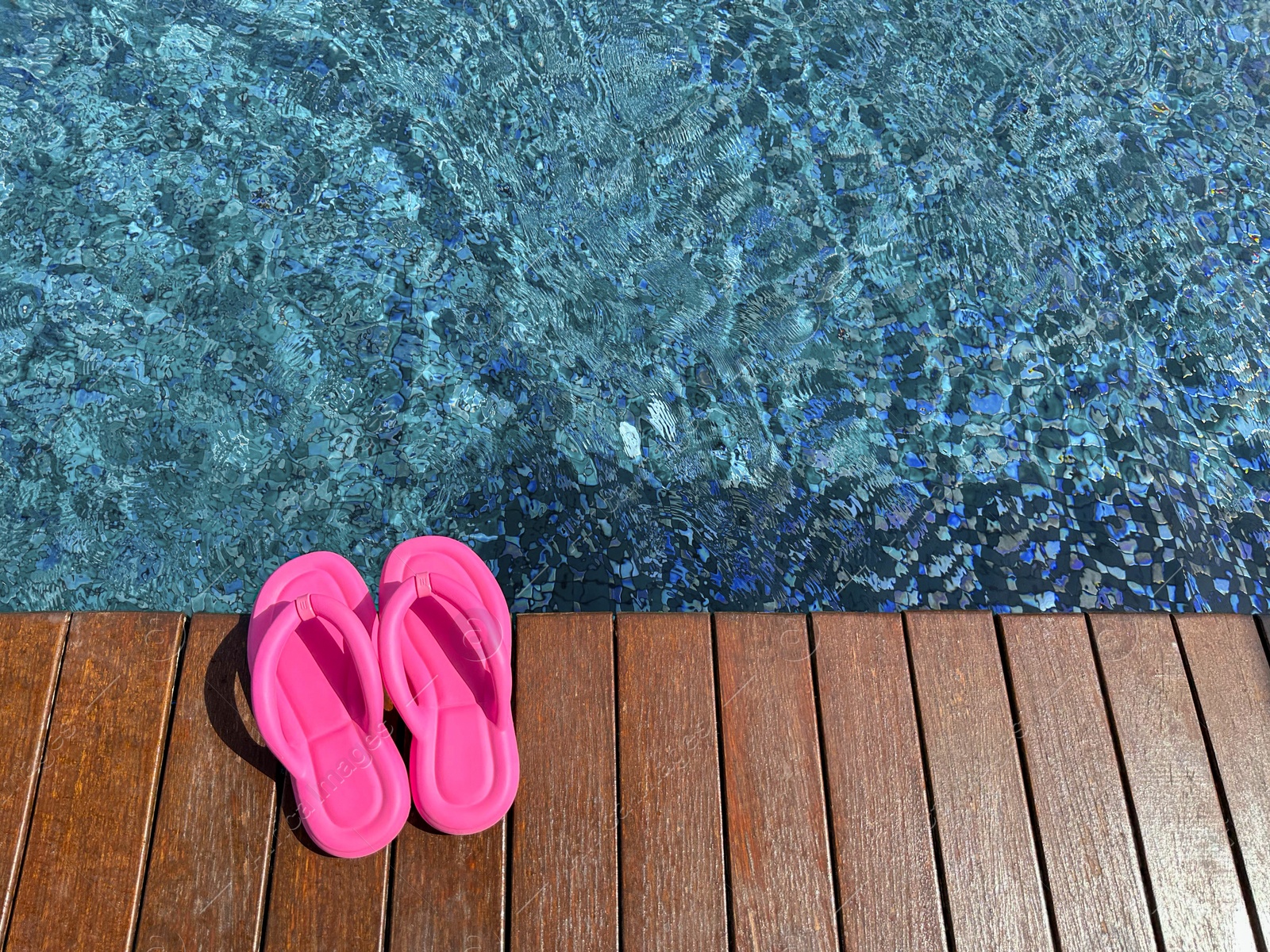 Photo of Clear rippled water in swimming pool and pink flip-flops on wooden deck outdoors, above view. Space for text
