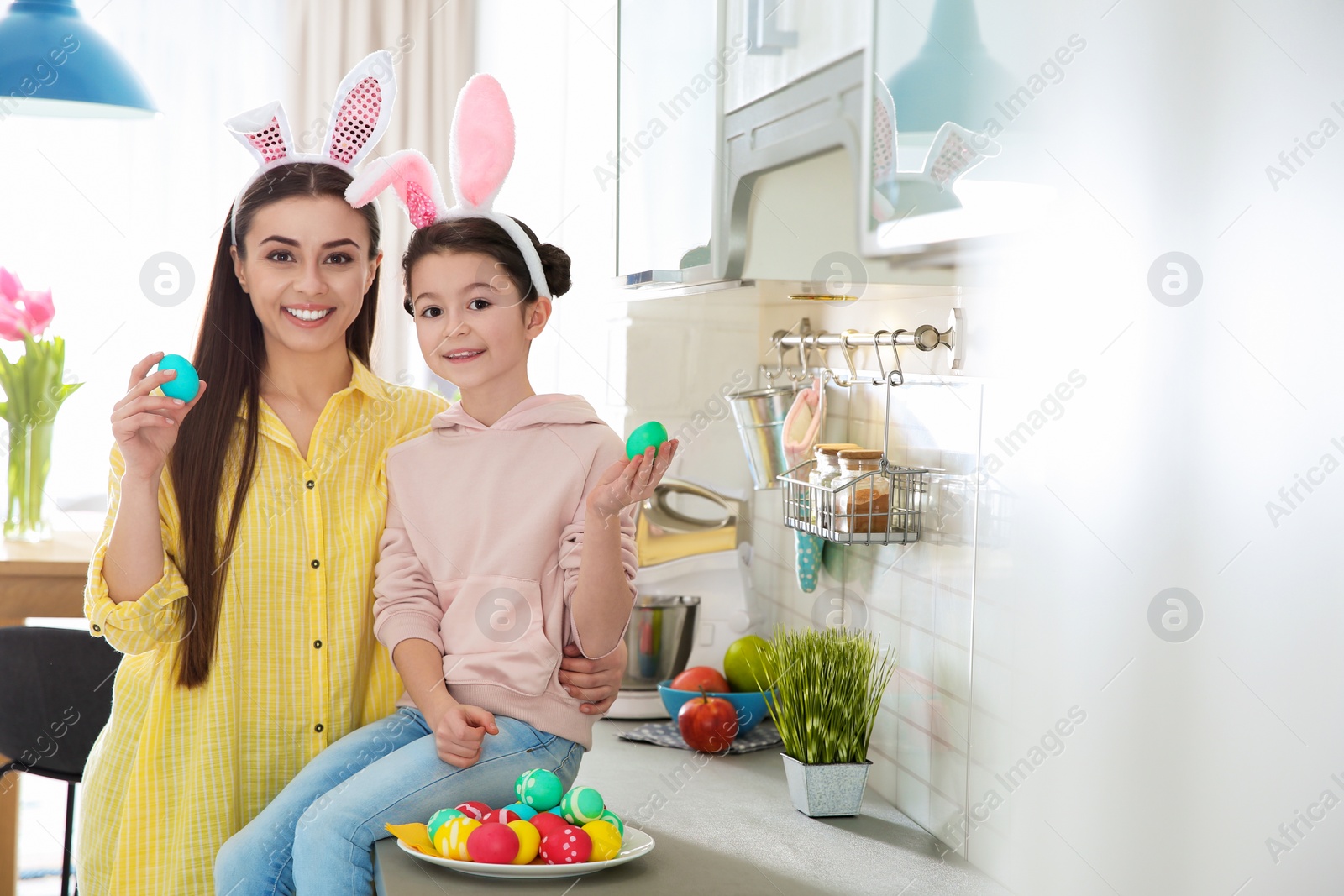 Photo of Mother and daughter with bunny ears headbands and painted Easter eggs in kitchen, space for text
