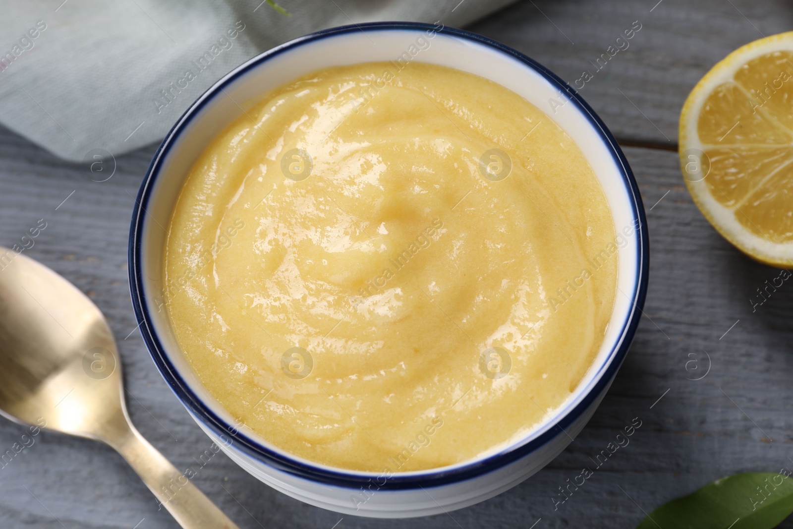 Photo of Delicious lemon curd in bowl, fresh citrus fruit and spoon on grey wooden table, closeup