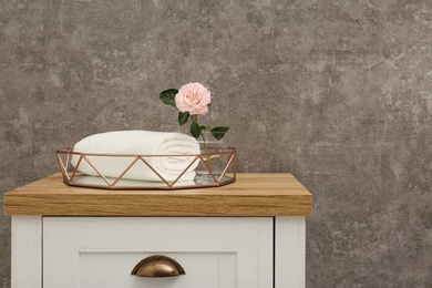 Photo of Rolled soft towel with beautiful flower in decorative tray on chest of drawers. Space for text