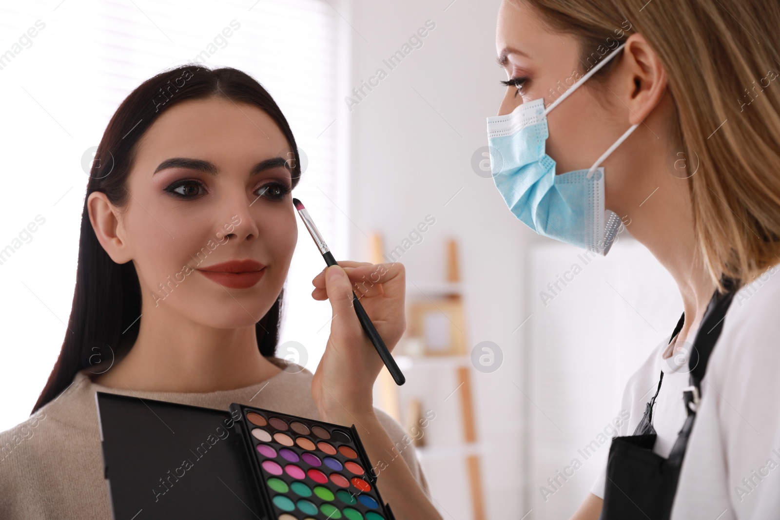 Photo of Makeup artist in protective mask working with woman indoors. Preventive measures during COVID-19 pandemic