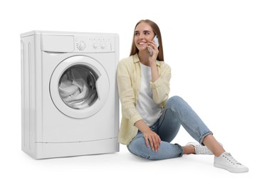 Beautiful young woman talking on smartphone near washing machine with laundry against white background