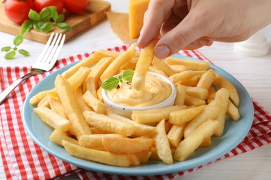 Photo of Woman dipping delicious French fries into cheese sauce at white table, closeup