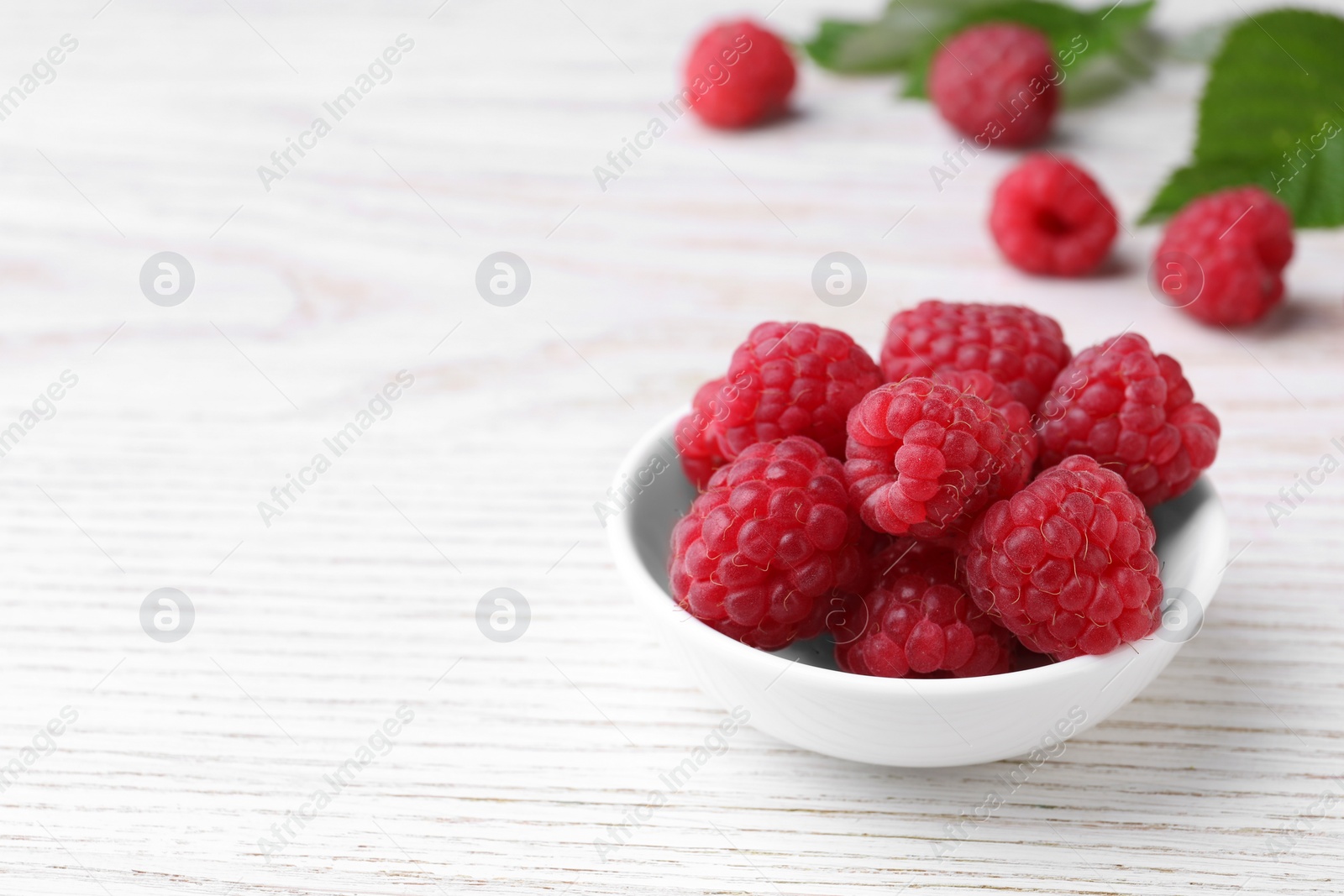 Photo of Tasty ripe raspberries in bowl on white wooden table, space for text