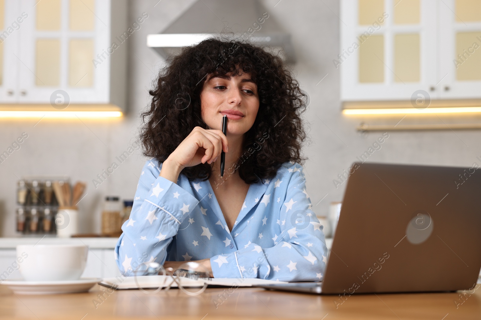 Photo of Beautiful young woman in stylish pyjama with pen using laptop at wooden table in kitchen
