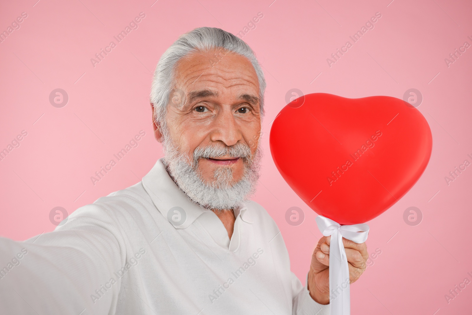 Photo of Senior man with red heart shaped balloon taking selfie on pink background