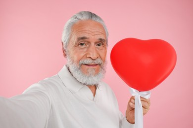Senior man with red heart shaped balloon taking selfie on pink background