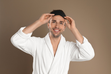 Photo of Happy young man in bathrobe and eye sleeping mask on brown background