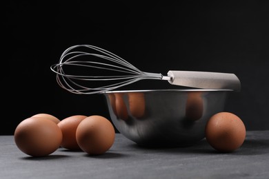 Metal whisk, raw eggs and bowl on dark grey table