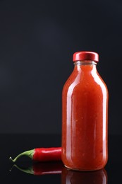 Photo of Spicy chili sauce in bottle and pepper against dark background, space for text