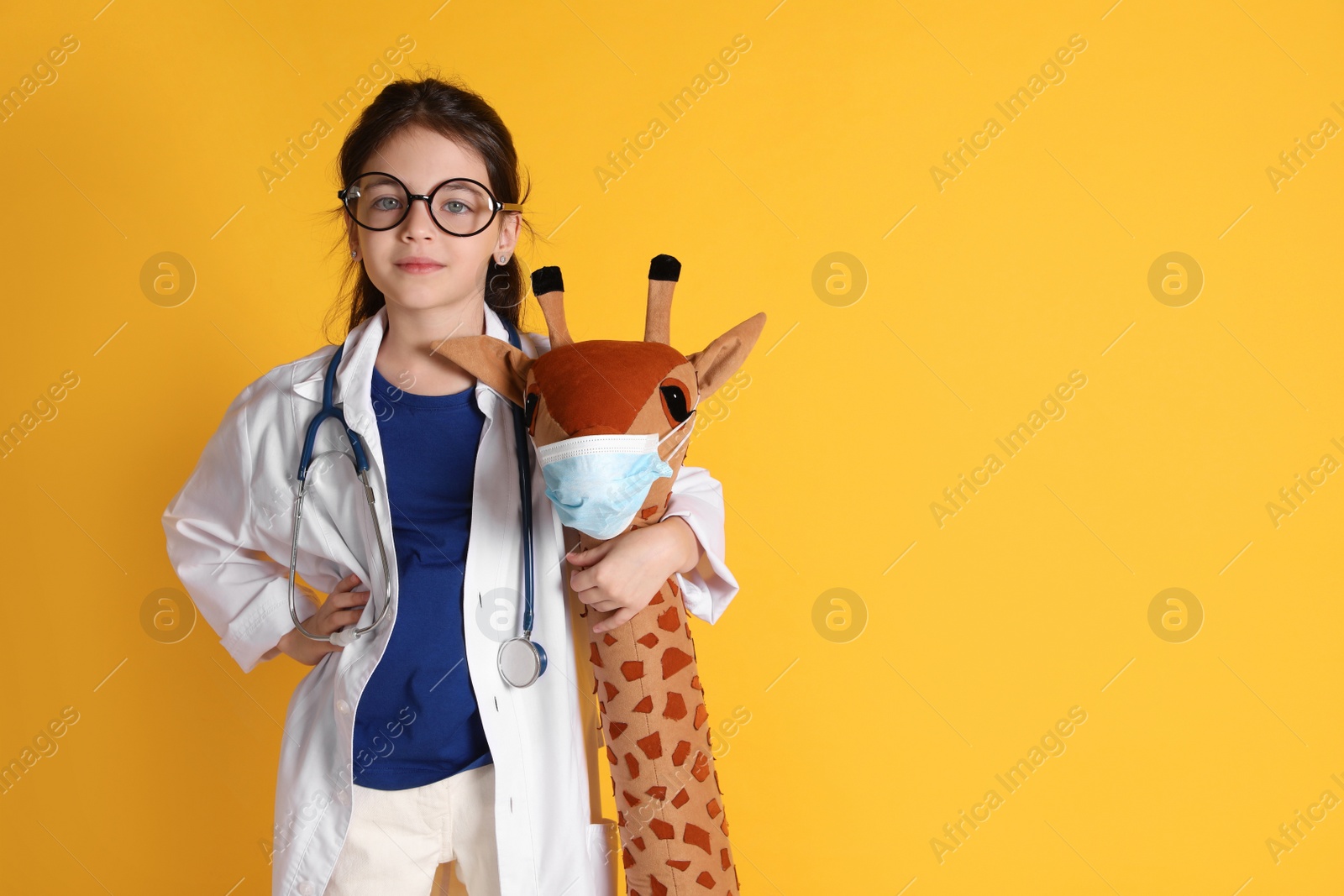 Photo of Little girl with eyeglasses, stethoscope dressed as doctor hugging toy giraffe on yellow background, space for text. Pediatrician practice