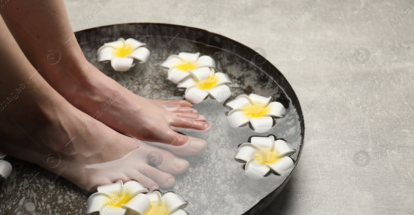 Photo of Woman soaking her feet in bowl with water and flowers on light grey floor, closeup with space for text. Spa treatment