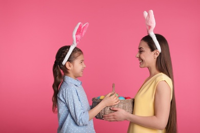 Photo of Mother and daughter in bunny ears headbands holding basket with Easter eggs on color background
