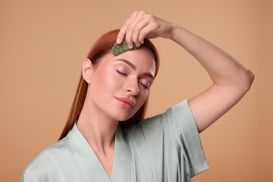 Photo of Young woman massaging her face with jade gua sha tool on pale orange background
