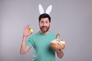 Portrait of happy man in cute bunny ears headband holding wicker basket with Easter eggs on light grey background