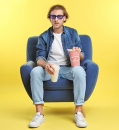 Emotional man with 3D glasses, popcorn and beverage sitting in armchair during cinema show on color background