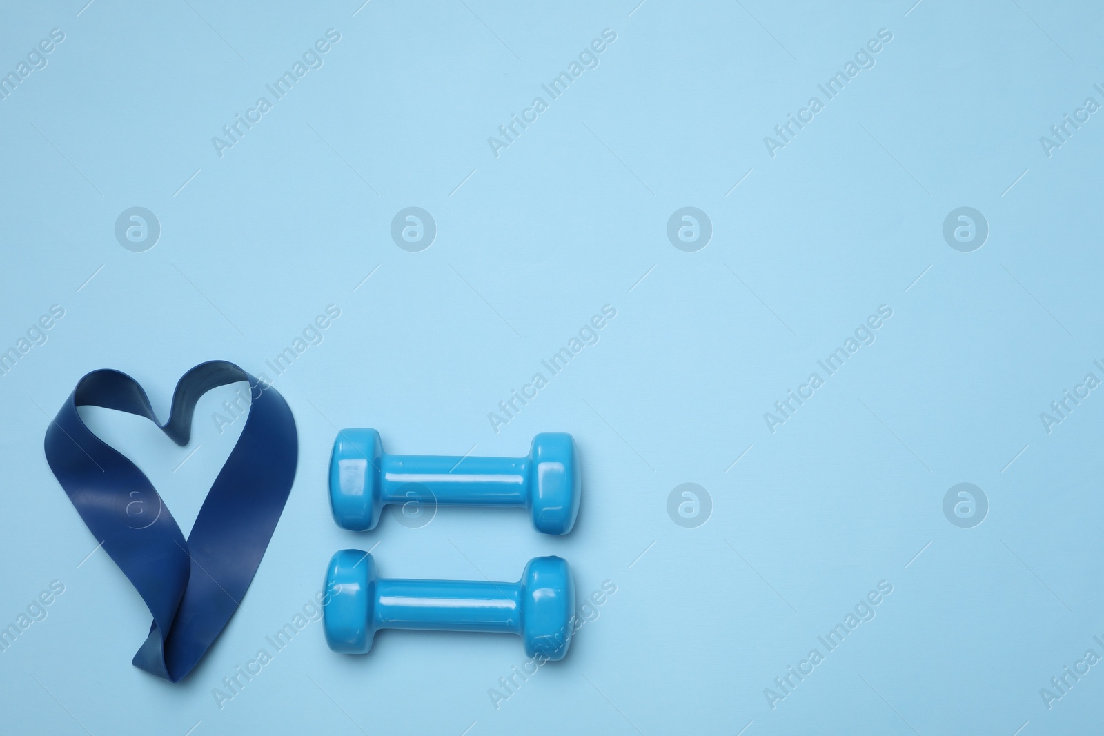 Photo of Two dumbbells and fitness elastic band in shape of heart on light blue background, flat lay. Space for text