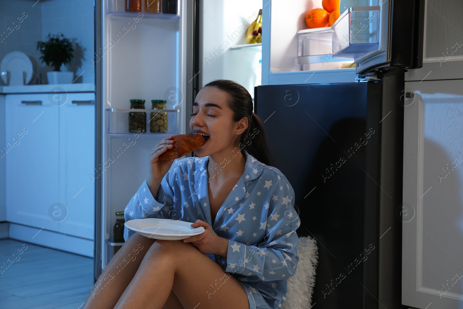 Photo of Young woman eating bun near open refrigerator at night