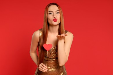 Photo of Young woman in dress with paper heart blowing kiss on red background