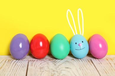 Image of Blue egg with drawn face and ears as Easter bunny among others on white wooden table against yellow background