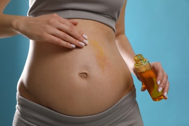 Pregnant woman applying cosmetic product on belly against light blue background, closeup