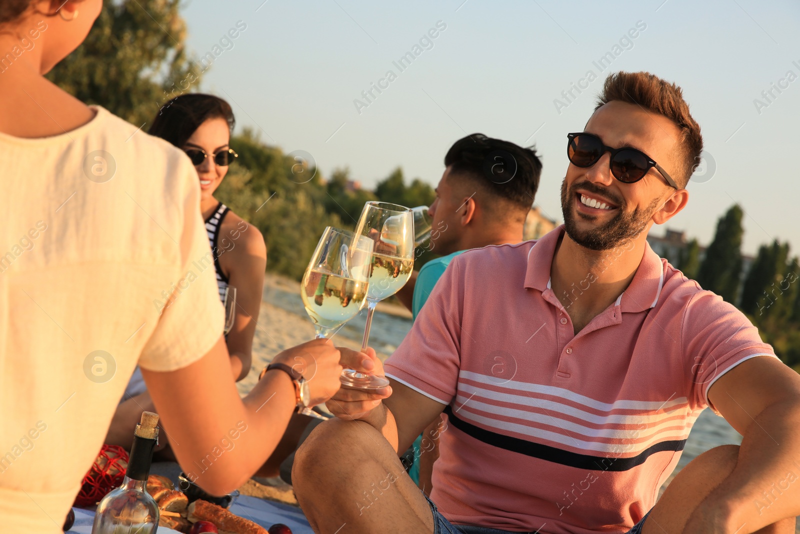 Photo of Group of friends having picnic outdoors at sunset, focus on glasses