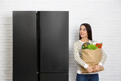 Young woman with paper bag full of products near refrigerator indoors