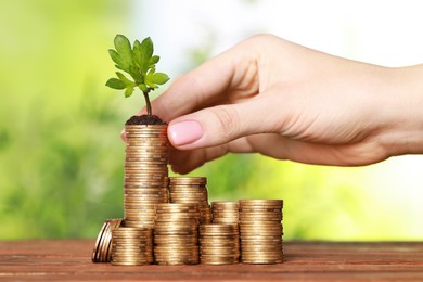 Woman putting coin with green sprout onto stack at wooden table against blurred background, closeup. Investment concept