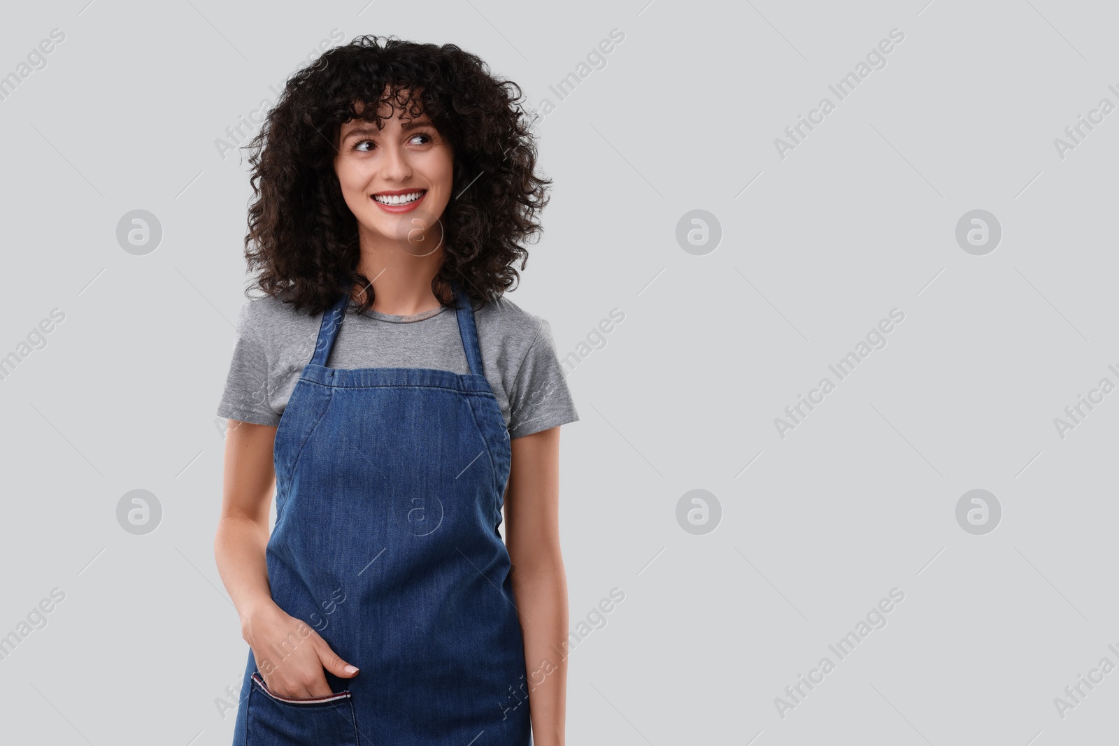 Photo of Happy woman wearing kitchen apron on light grey background, space for text. Mockup for design