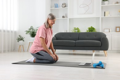 Senior woman in sportswear on fitness mat near laptop at home