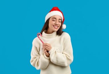 Photo of Young woman in beige sweater and Santa hat holding candy cane on light blue background. Celebrating Christmas