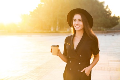 Photo of Beautiful young woman in stylish black dress and hat with cup of coffee on city street