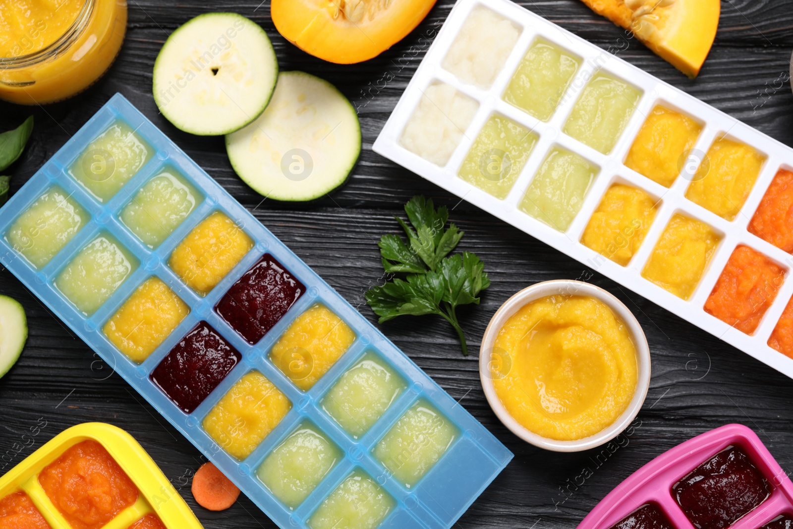 Photo of Different frozen purees in ice cube trays and ingredients of black wooden table, flat lay. Ready for freezing