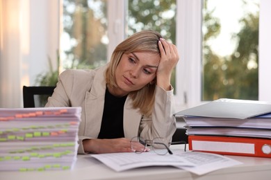 Photo of Overwhelmed woman sitting at table with stacks of documents and folders in office