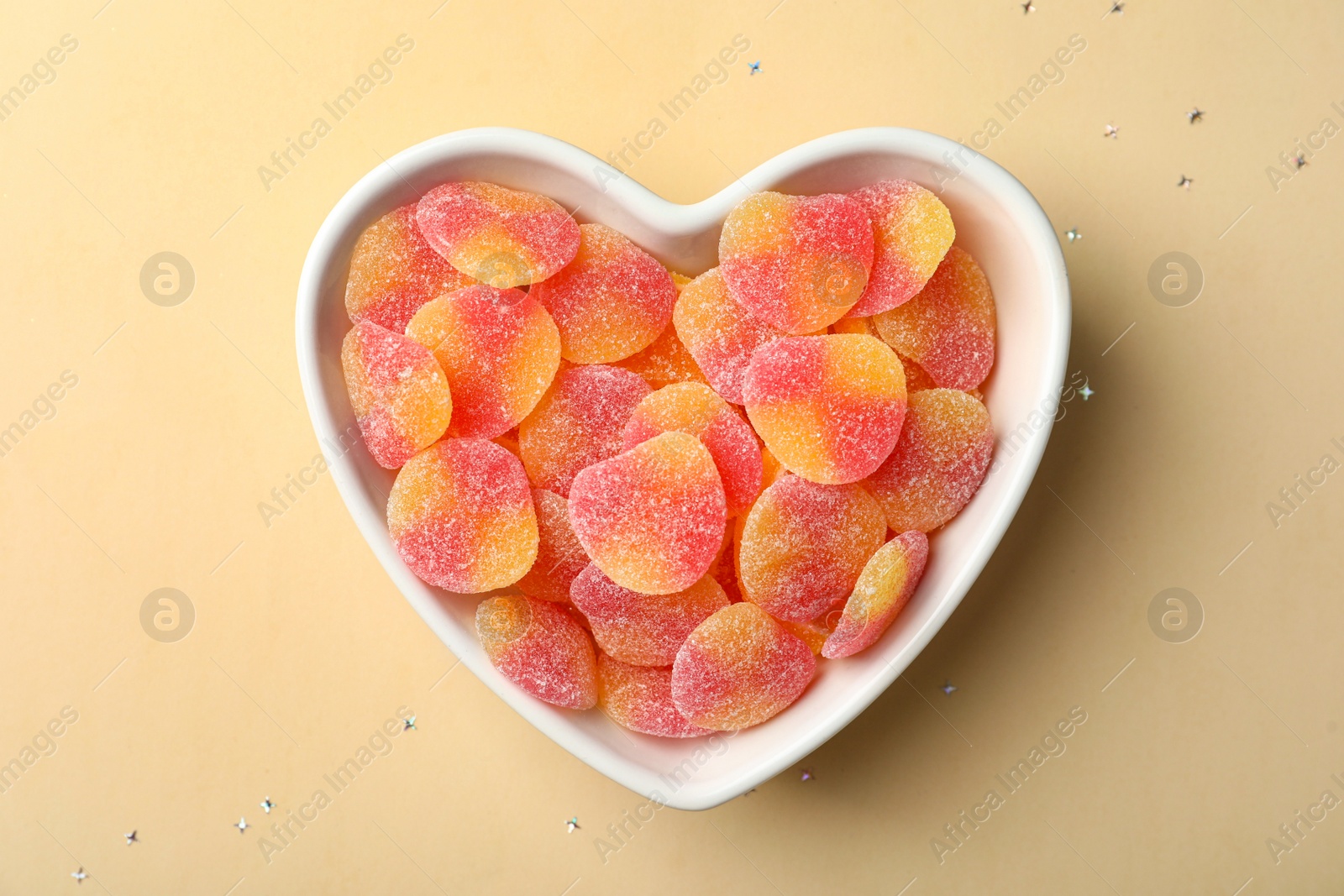 Photo of Tasty jelly candies in heart shaped bowl on beige background, top view