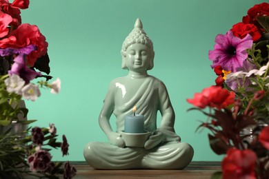 Photo of Buddhism religion. Decorative Buddha statue with burning candle and beautiful flowers on table against turquoise wall, selective focus