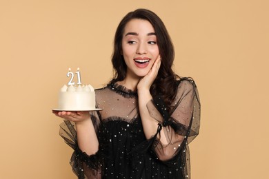 Photo of Coming of age party - 21st birthday. Surprised woman holding delicious cake with number shaped candles on beige background
