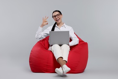 Photo of Happy woman with laptop sitting on beanbag chair and showing okay gesture against light gray background