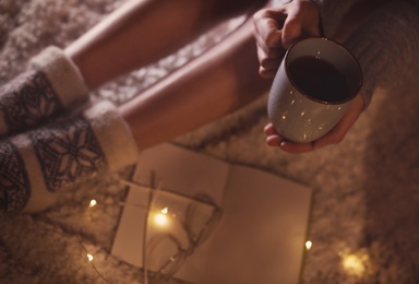 Woman with cup of hot beverage and book at home in winter evening, closeup