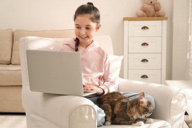 Cute little girl with laptop and cat in armchair at home. First pet