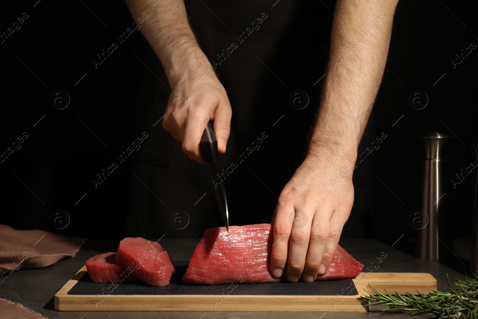 Photo of Man cutting fresh raw meat on table against dark background, closeup