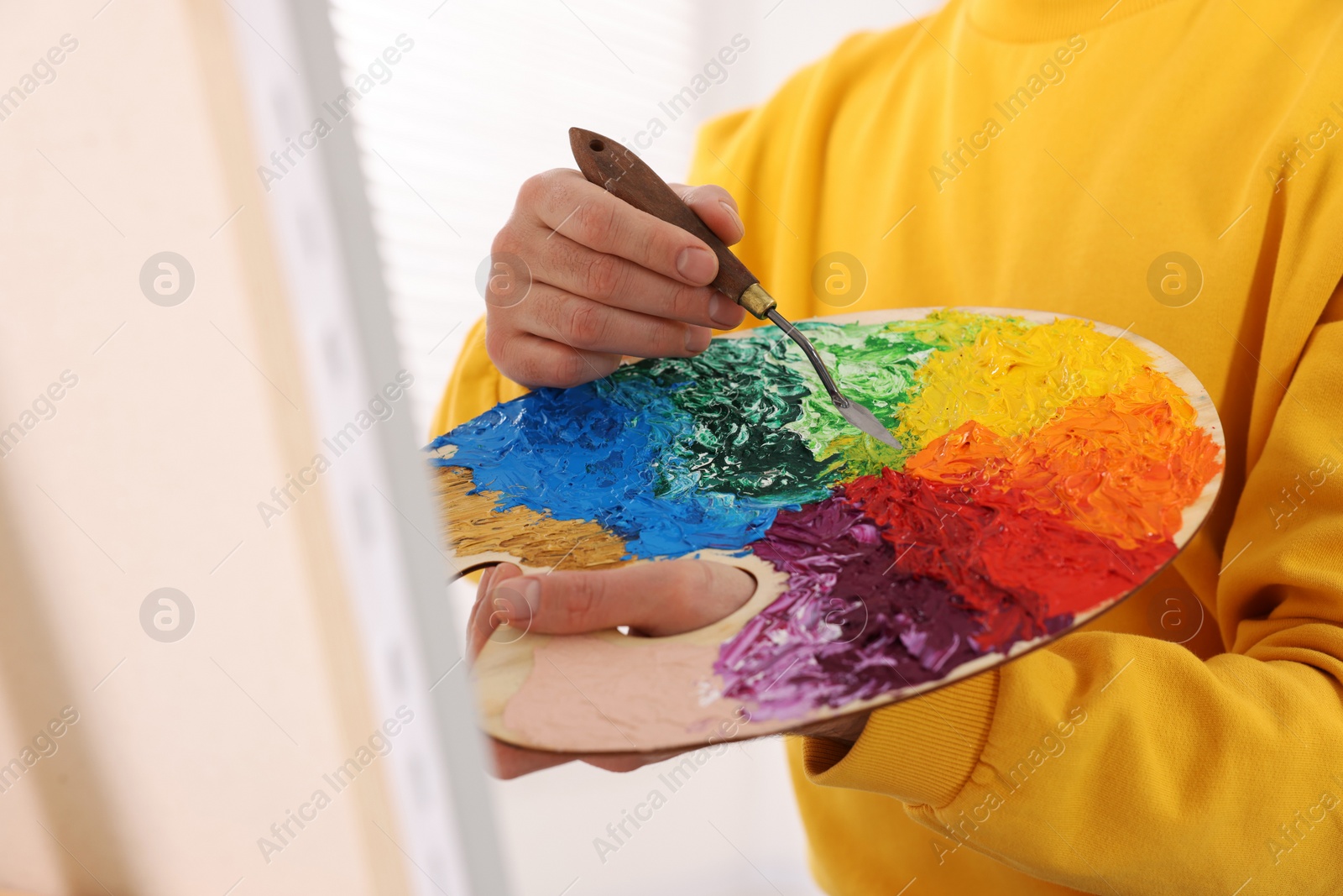 Photo of Man mixing paints on palette with brush near canvas in studio, closeup