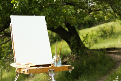 Photo of Easel with blank canvas and painting equipment in picturesque countryside