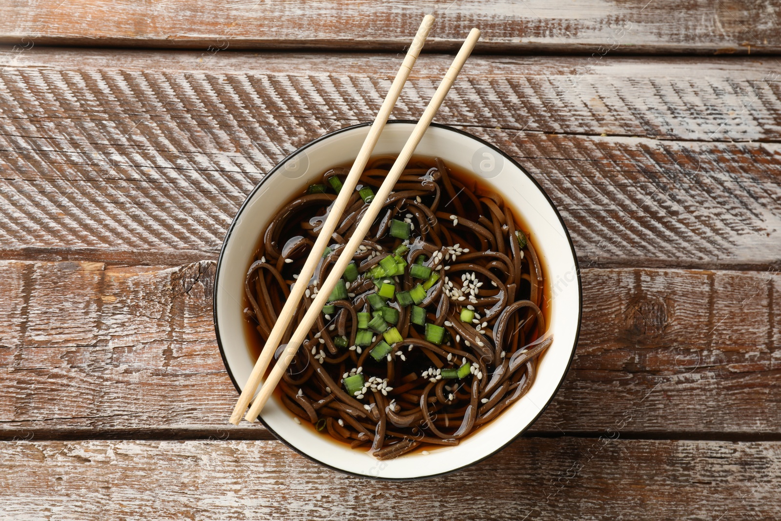 Photo of Tasty soup with buckwheat noodles (soba), onion in bowl and chopsticks on wooden table, top view