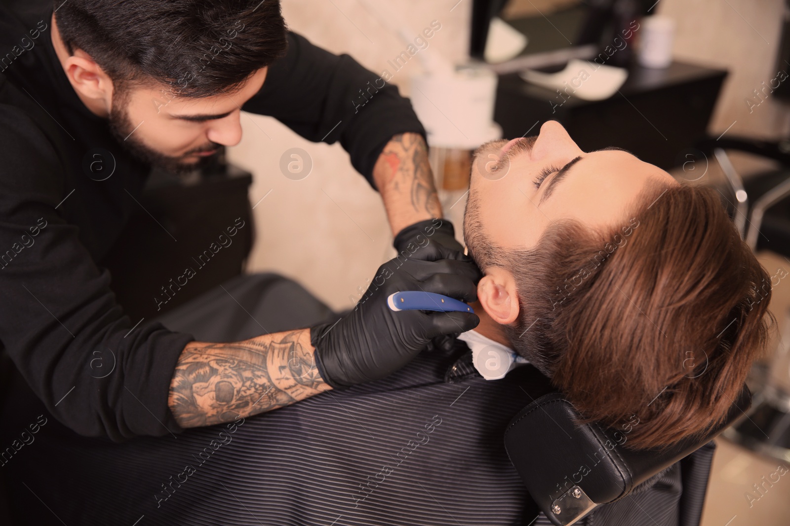 Photo of Professional hairdresser shaving client with straight razor in barbershop