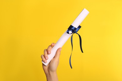 Student holding rolled diploma with blue ribbon on yellow background, closeup